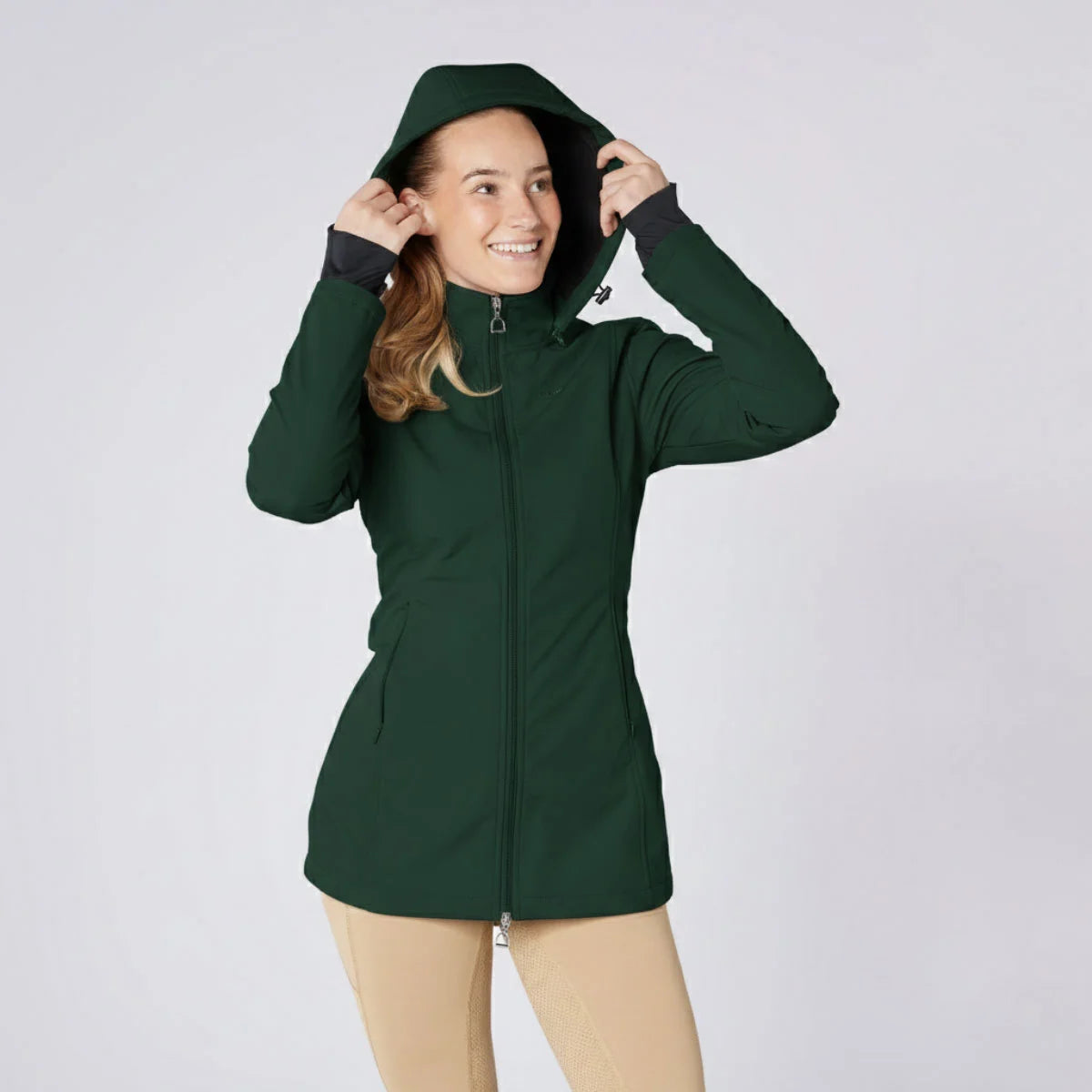 CAMEO Zenith Pro Ride Coat in EverGreen modelled with the storm hood up and paired with beige breeches, showcasing the waterproof longline cut, high fleece collar, two-way stirrup zip, and thumb-loop cuffs engineered for cold-weather riding and yard protection.
