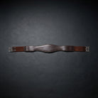 A brown leather horse girth with silver metal buckles, displayed horizontally on a dark textured background.